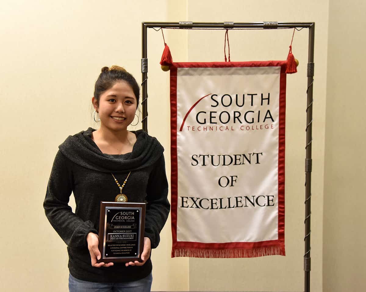 Kanna Suzuki, October 2017 Student of Excellence, stands in front of the Student of Excellence banner while holding her plaque.