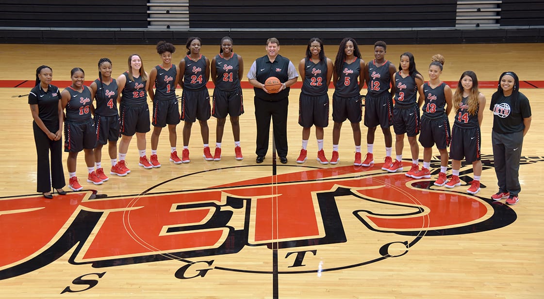 The 2017-2018 Lady Jets basketball team stands in a semi-circle on the mid-court line of a basketball court. Standing left to right are: Assistant Coach Kezia Conyers, CeDeja James (10), Kayla Holmes (4), Aubrey Maulden (20), Ricka Jackson (1), Esther Adenike (24), Bigue Sarr (21), Coach James Frey, Sceret Ethridge (22), Houlfat Mahouchia (5), Fatou Pouye (12), Dakata Toney (3), Alyssa Nieves (13), Kanna Suzuki (14), Student Assistant Davesha Murray.