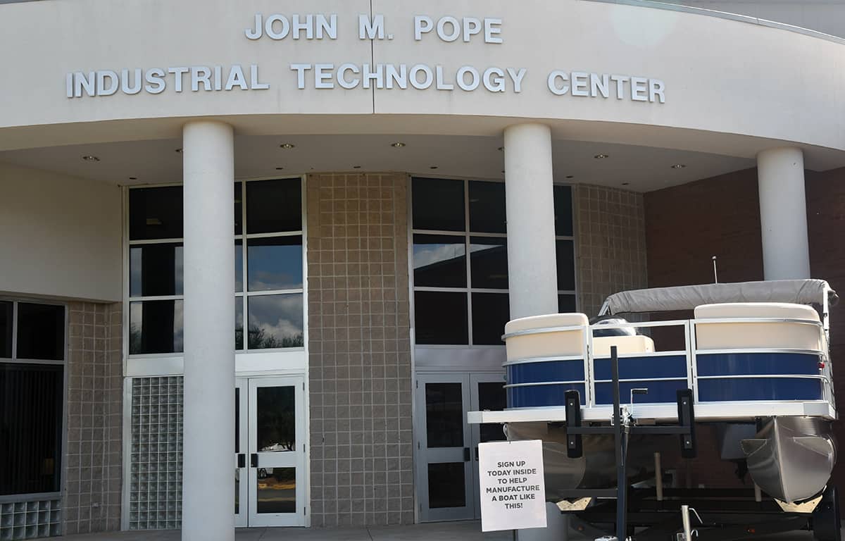 A pontoon boat sits at an angle in front of the John M. Pope Technology Center on the Americus campus of South Georgia Technical College. A sign reads "Sign Up Today Inside To Help Manufacture A Boat Like This!"