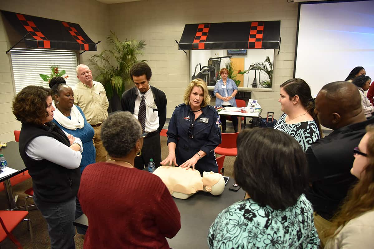 A group of SGTC employees stand in a circle around a CPR training dummy while a woman (Monica Hood) gives instruction
