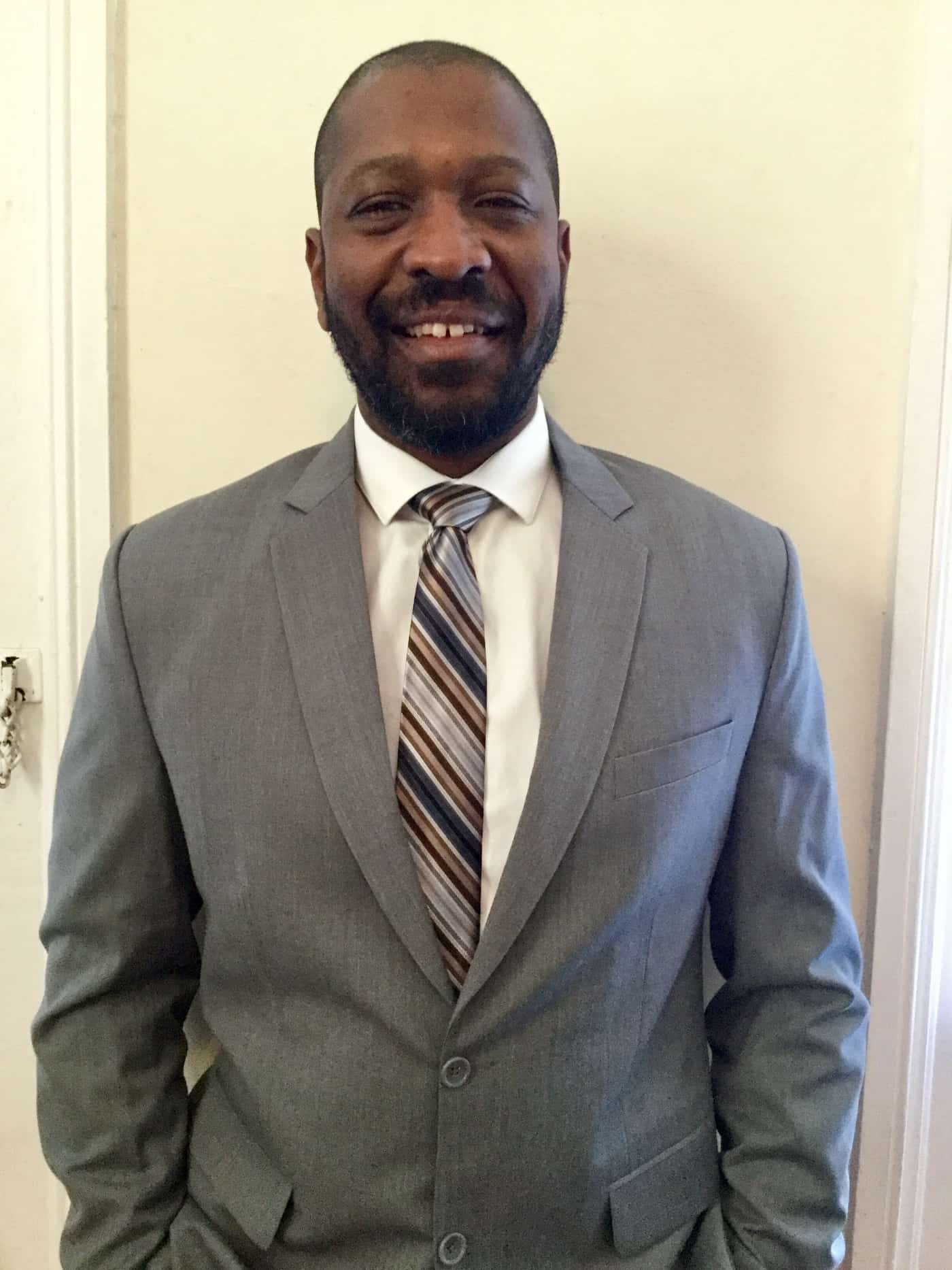 Image of Ronny Roundtree, Jr. standing against a wall and dressed in a gray suit.