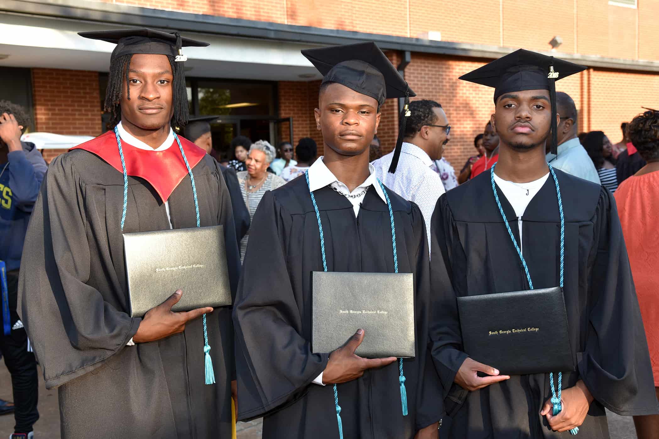 Three males stand together in their black graduation caps and gowns holding their diplomas in front of them. They each wear turquoise cords around their necks.