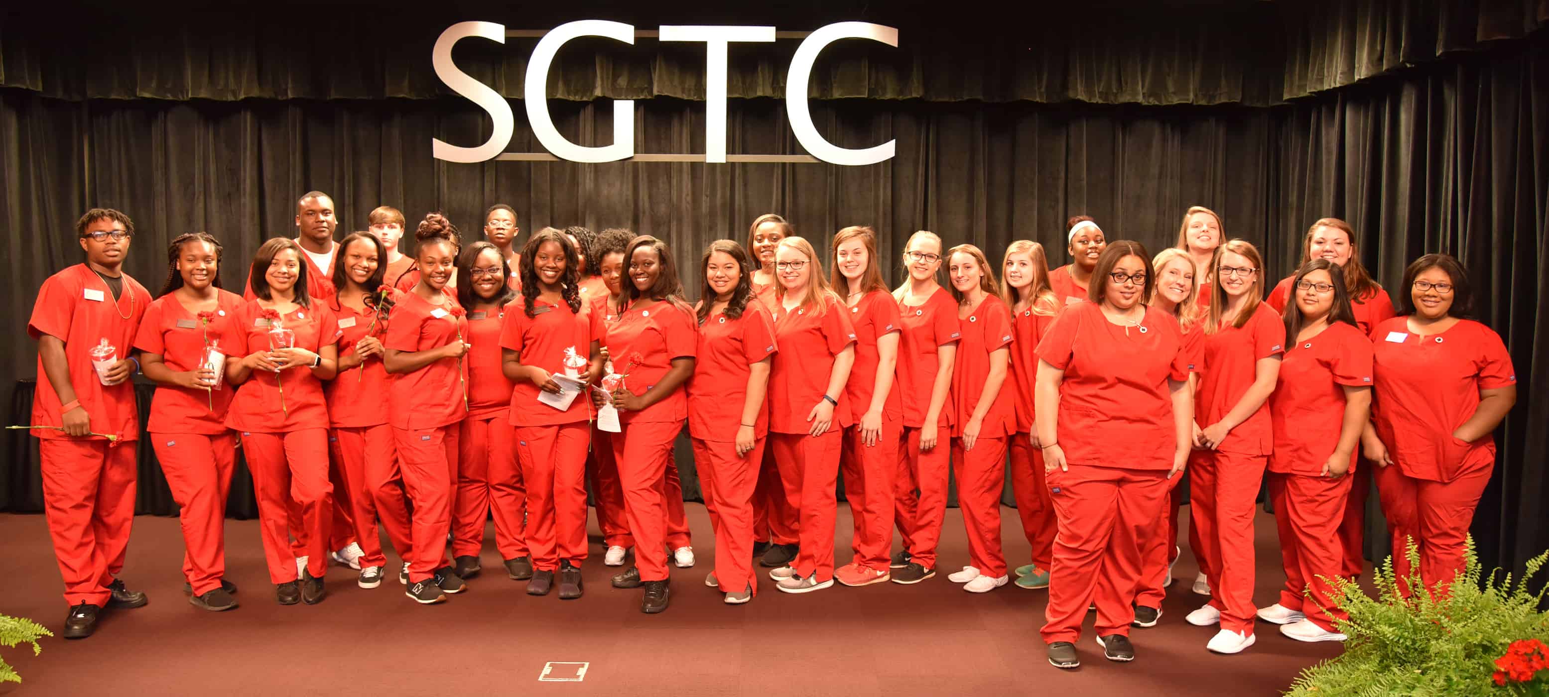 29 high school students stand on a stage wearing red scrubs.