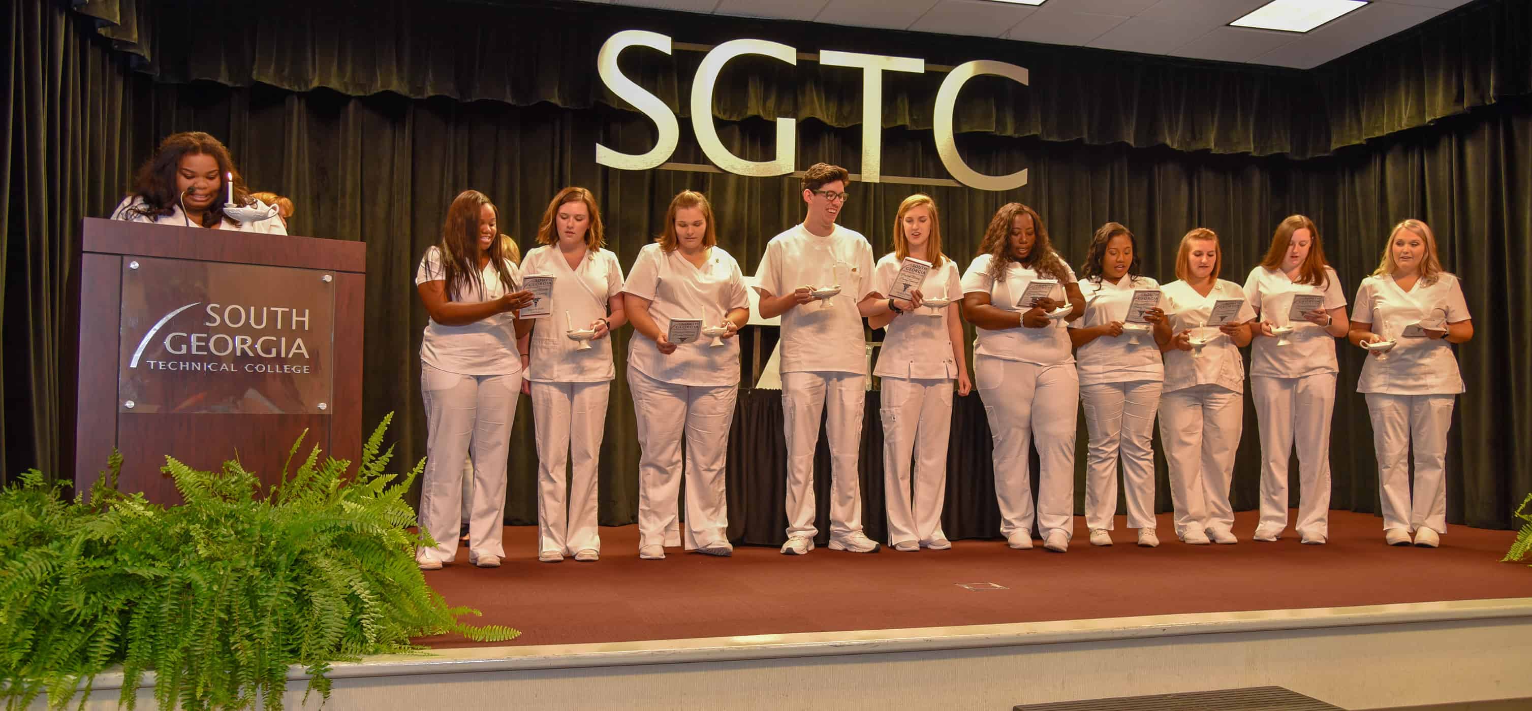 11 people stand on a stage wearing white scrubs, holding candles.