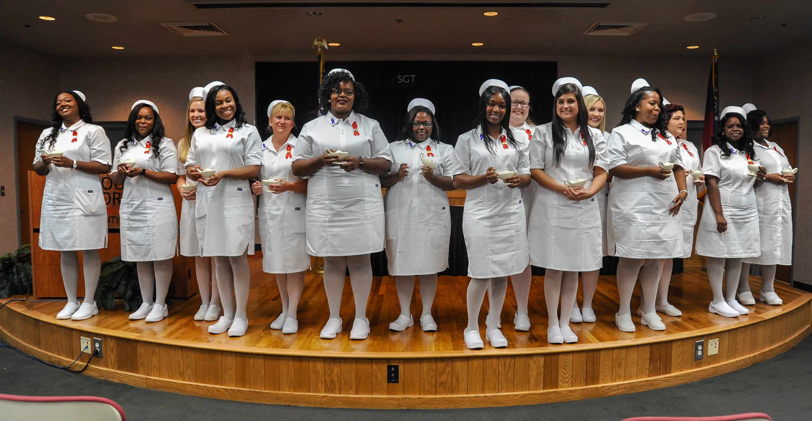 A group of 15 females in white nurse dresses stand side by side on a stage, holding candles.