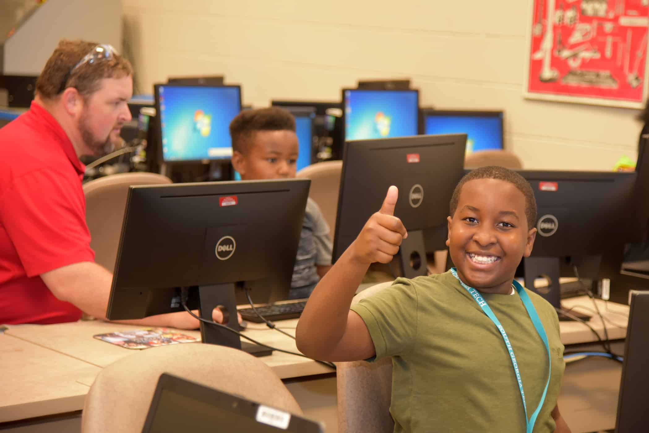 Kaylon Harvey, a seventh grader from Sumter County Middle School, gives a thumbs up during a session of the recent STEM camp while precision machining and manufacturing instructor Chad Brown helps a student design a 3D model in the background.
