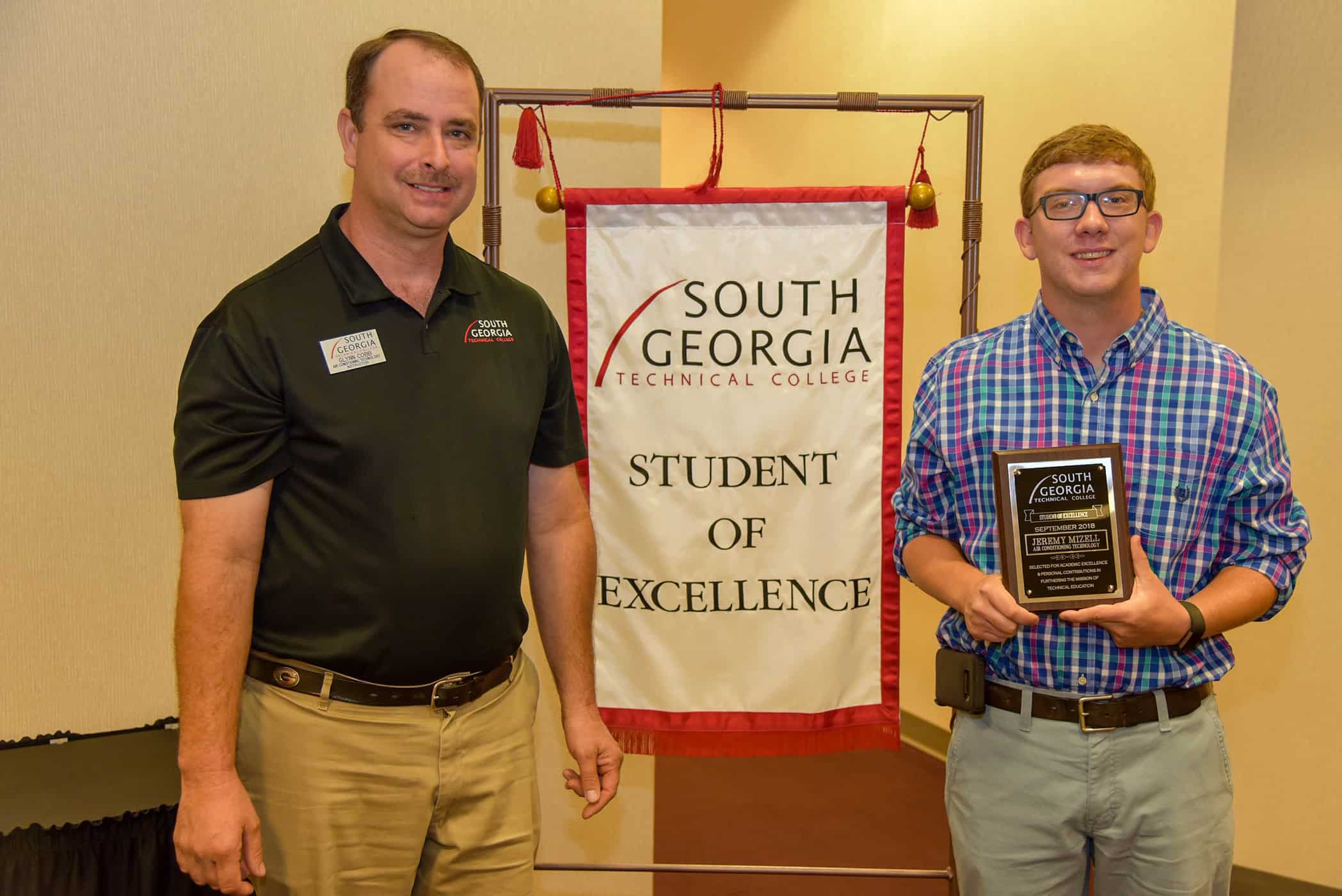 Two men stand in front of a white banner that reads "Student of Excellence"