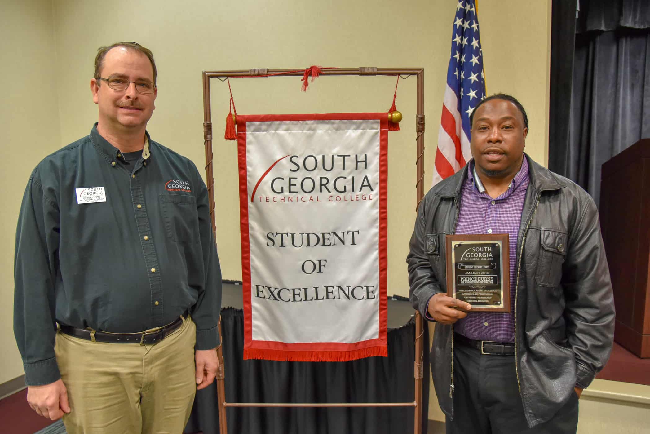 Two men stand together on either side of a banner that reads "Student of Excellence"