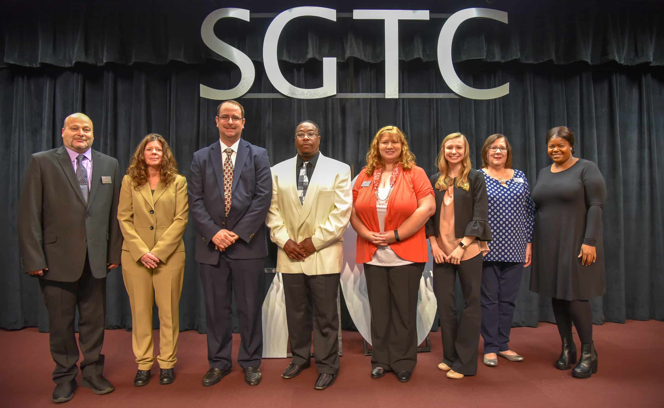Eight adults stand on a stage , side by side.