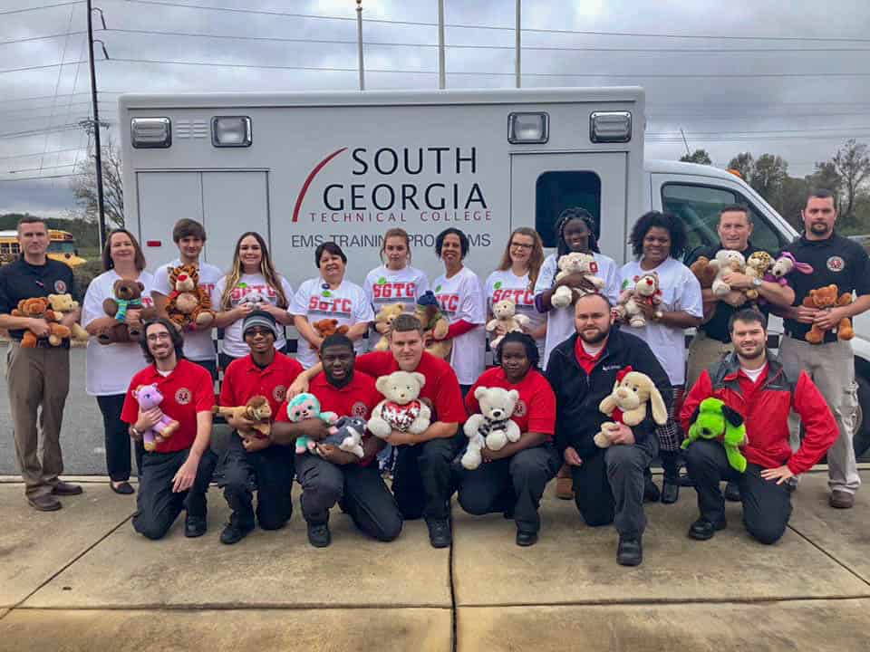 A group of people stand in front of an ambulance holding stuffed animals.