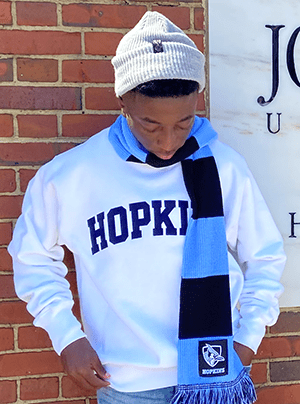 SGTC CIS Alumnus Chance Simpson is shown above in front of the Johns Hopkins University sign and looking at his Johns Hopkins scarf and sweatshirt.